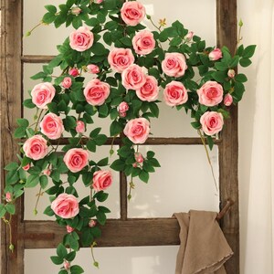 May include: A wooden window frame adorned with climbing vines of pink roses and green leaves. The roses are in full bloom, with a soft, romantic aesthetic. A tan cloth hangs from the window's handle.