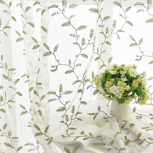 May include: White sheer curtains with an embroidered green leaf pattern. The curtains are hanging in a window and there is a vase of white flowers on a table in front of them.
