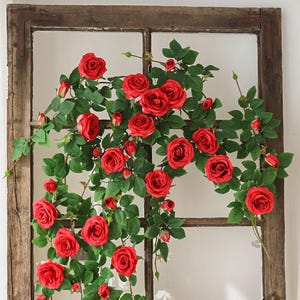May include: A climbing vine with vibrant red roses and green leaves, arranged on a rustic, wooden window frame. The roses are in full bloom, creating a visually appealing floral display. The window frame has a weathered appearance.
