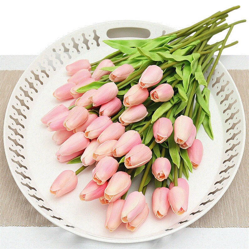 Op de afbeelding: Een boeket van roze kunstmatige tulpen met groene stelen en bladeren, gerangschikt op een witte dienblad met een decoratieve rand.