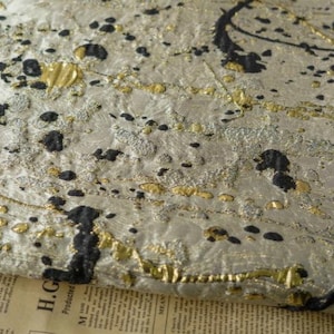 Pode incluir: Close-up de um tecido prateado e dourado com um design de respingos de tinta preta. O tecido tem um aspecto texturizado e tecido.