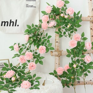 May include: A long garland of artificial pink roses with green leaves, draped over a wooden ladder. The garland is perfect for adding a touch of spring to any home.