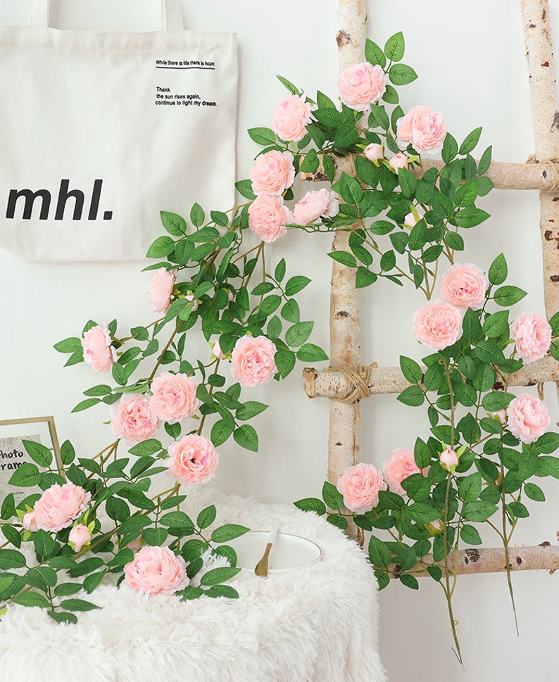 May include: A long garland of artificial pink roses with green leaves, draped over a wooden ladder. The garland is perfect for adding a touch of spring to any home.