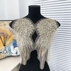 May include: A pair of gold angel wings with sequins and pearl embellishments. The wings are attached to a mesh fabric backing.