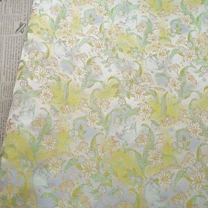 May include: A piece of fabric featuring a repeating floral pattern with white lily-like flowers, light green leaves, and soft yellow watercolor-style washes. The textile is displayed next to an open book with vintage newspaper-style text.