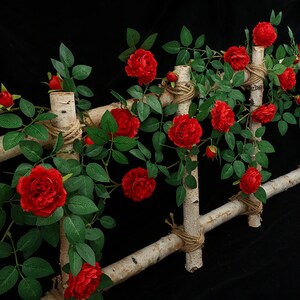 May include: Artificial red rose vine with green leaves climbing a wooden trellis. The vine is decorated with twine and is perfect for adding a touch of nature to any space.
