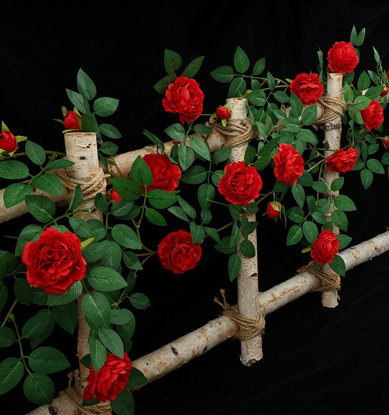 May include: Artificial red rose vine with green leaves climbing a wooden trellis. The vine is decorated with twine and is perfect for adding a touch of nature to any space.