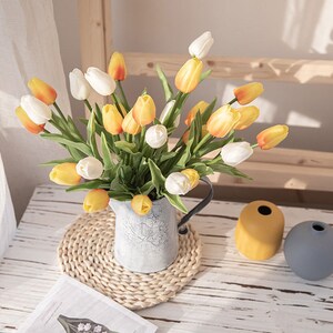 Op de afbeelding: Een boeket gele en witte tulpen gerangschikt in een rustieke metalen kan. De kan staat op een geweven placemat op een houten tafel. Er zijn twee decoratieve vazen op de achtergrond.
