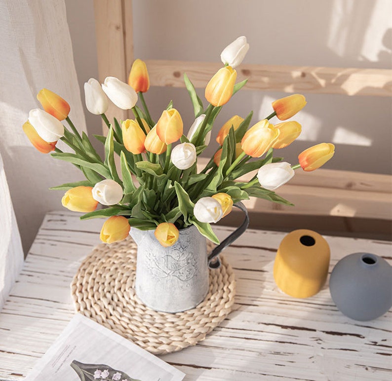 Op de afbeelding: Een boeket gele en witte tulpen gerangschikt in een rustieke metalen kan. De kan staat op een geweven placemat op een houten tafel. Er zijn twee decoratieve vazen op de achtergrond.
