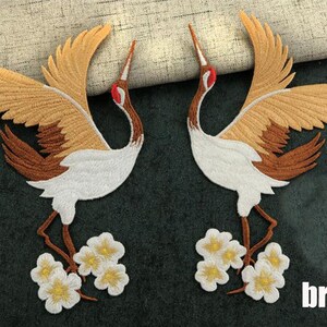May include: Two brown and white embroidered crane appliques with white flowers. The cranes are facing each other with their wings spread. The flowers are at the bottom of the cranes' legs.