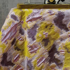 Puede incluir: Una tela texturizada con un patrón abstracto de manchas en amarillo mostaza, malva metálico y morado oscuro. El material tiene una superficie tejida en relieve con un brillo distintivo, drapeada sobre una superficie con una base de madera visible.