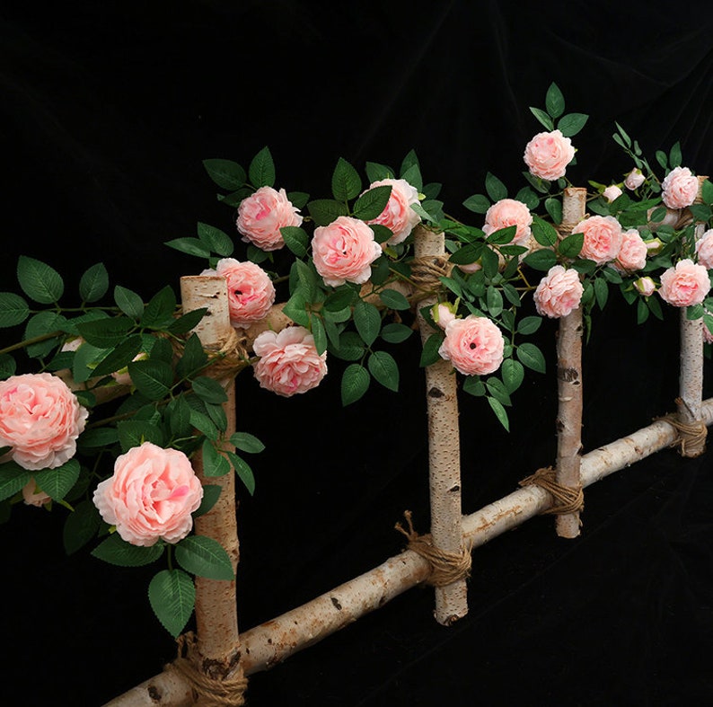 May include: A pink artificial rose vine with green leaves is draped over a wooden trellis. The trellis is made of natural birch branches and is tied together with twine.