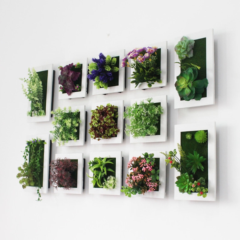 Puede incluir: Un conjunto de 12 piezas de decoraci&oacute;n de pared con marco blanco que presentan plantas y flores artificiales. Los marcos est&aacute;n dispuestos en un patr&oacute;n de cuadr&iacute;cula en una pared blanca. Las plantas y las flores son de varios tonos de verde, morado y rosa.