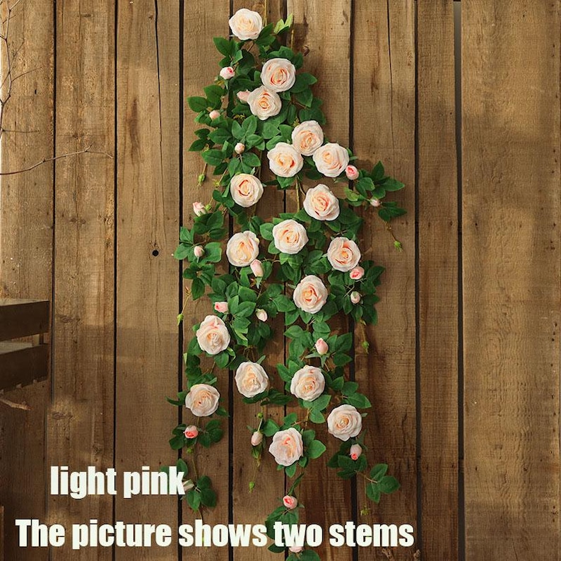 May include: Two stems of light pink artificial roses with green leaves cascade down a wooden background. The image shows the text "light pink" and "The picture shows two stems".