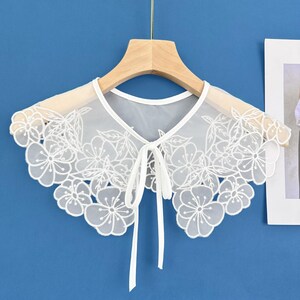 May include: White lace collar with a floral design and a bow tie. The collar is on a wooden hanger against a blue background.