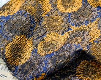 May include: Close-up of a textured fabric with a floral pattern. The fabric features a combination of gold, navy blue, and dark gray colors. The design includes large, stylized flower shapes, creating a rich and detailed textile.