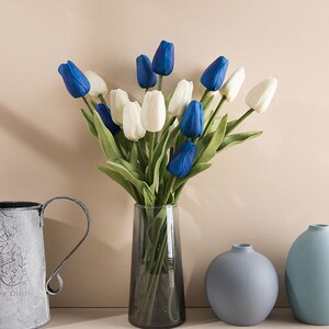 Op de afbeelding: Een boeket kunstmatige tulpen in een glazen vaas. De tulpen zijn wit en blauw, en de vaas staat op een tafel met twee kleinere blauwe vazen.