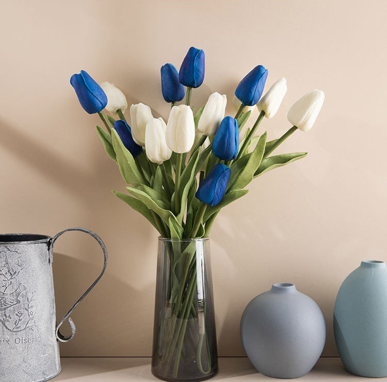 Op de afbeelding: Een boeket kunstmatige tulpen in een glazen vaas. De tulpen zijn wit en blauw, en de vaas staat op een tafel met twee kleinere blauwe vazen.
