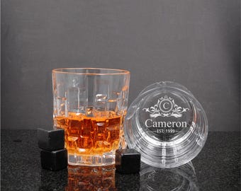 May include: Two clear glass tumblers with a textured geometric base. One glass is filled with amber liquid and three black square stones. The other glass is empty and engraved with a crest, the name Cameron, and the text EST: 1999.