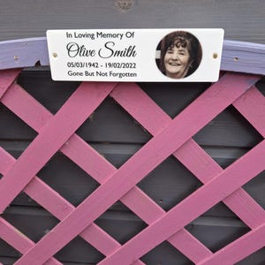 May include: A white memorial plaque with the text "In Loving Memory Of Olive Smith 05/03/1942 - 19/02/2022 Gone But Not Forgotten" and a photo of a woman. The plaque is mounted on a pink lattice fence.