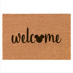May include: A brown coir doormat with the word "welcome" in black cursive lettering and a black Mickey Mouse silhouette in the middle of the word.