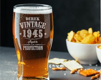 Może przedstawiać: Szklanka do piwa z przezroczystego szkła wypełniona bursztynowym płynem i pianą, z wygrawerowanym napisem DEREK VINTAGE 1945 Aged to PERFECTION. Szklanka ma krople kondensacji na zewnętrznej powierzchni oraz szeroki, zwężający się cylindryczny kształt.