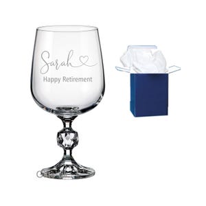 Può includere: Un calice in vetro trasparente con stelo sfaccettato, inciso con il nome Sarah, un simbolo a forma di cuore e il testo Happy Retirement. Accanto al bicchiere si trova una piccola scatola regalo blu scuro aperta contenente carta velina bianca.