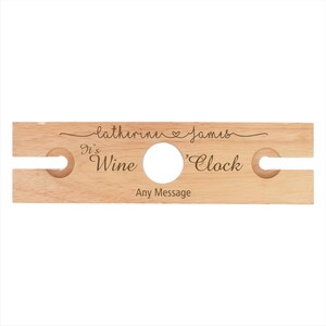 May include: Personalized wooden wine bottle holder with two slots for glasses. It reads "It's Wine 'Clock" and "Catherine & James" with a heart symbol.