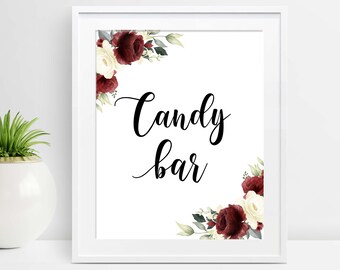 May include: A framed white sign with the words "Candy bar" in elegant black script. The sign is decorated with watercolor floral arrangements in burgundy, cream, and green, perfect for weddings or events.