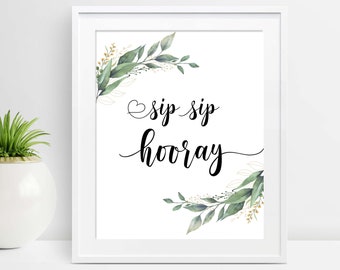 May include: White framed art print with the words "sip sip hooray" in black script. The print is decorated with watercolor greenery and gold accents in the corners. A decorative plant in a white pot is visible in the background.