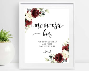 May include: White framed sign with the words "mom-osa bar" in elegant script. The sign includes instructions and the word "cheers!" Floral accents in burgundy and cream adorn the corners, creating a festive look.