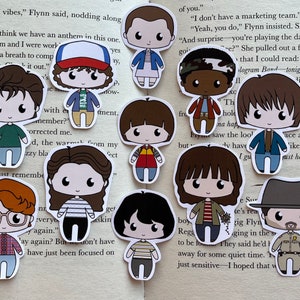May include: A collection of die-cut stickers featuring cartoon illustrations of characters from a popular science fiction series. The stickers depict characters with large eyes and simplified features, wearing outfits from the show. The stickers are on a book page.