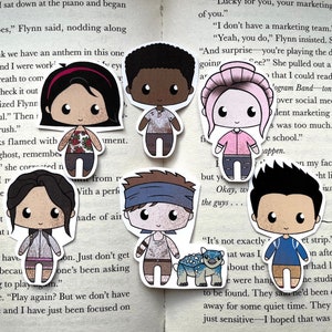 Puede incluir: Una colección de siete pegatinas de personajes estilo chibi, cada una con ojos grandes y atuendos únicos. Las pegatinas presentan una variedad de tonos de piel y peinados. Una pegatina incluye un pequeño dinosaurio azul. Las pegatinas están en una página de libro.