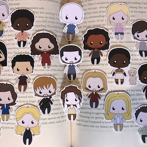 May include: A collection of die-cut stickers depicting cartoon characters with diverse skin tones, hair colours, and clothing. The stickers are arranged on the open pages of a book, creating a playful display. The characters have large eyes and simple facial features.