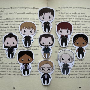 May include: A selection of cartoon stickers depicting characters in medical scrubs. Each sticker has a different hairstyle and skin tone, with a consistent design of black scrubs and a white coat. The stickers are arranged on a book page.
