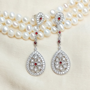 May include: A pair of silver teardrop earrings with a red stone in the center of each teardrop. The earrings are surrounded by clear stones and are hanging from a pearl necklace.
