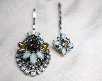 May include: Two silver hair clips with decorative embellishments. The larger clip features a black and gold centre with white and yellow stones. The smaller clip features white and blue stones.