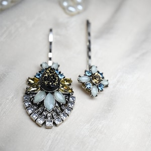 May include: Two silver hair clips with decorative embellishments. The larger clip features a black and gold center with white and yellow stones. The smaller clip features white and blue stones.