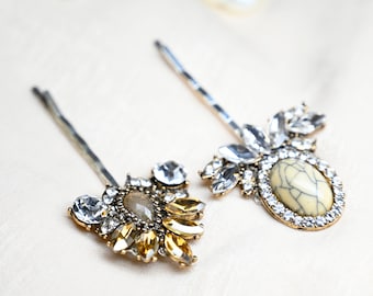 May include: Two decorative hair pins with jewel embellishments. One pin has clear and amber crystals, the other a cream-coloured oval stone surrounded by clear crystals. A pearl necklace is in the background. The pins are on a cream-coloured surface.