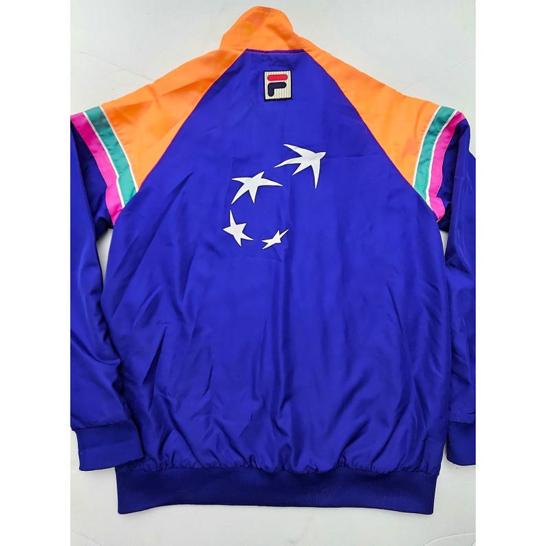 Peut inclure: Veste bleue vintage avec des accents orange et roses. La veste pr&eacute;sente un motif d'&eacute;toiles blanches au dos et le logo de la marque sur la poitrine. La veste a une fermeture &eacute;clair compl&egrave;te et un ourlet et des poignets c&ocirc;tel&eacute;s.