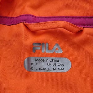 Peut inclure: Gros plan sur un v&ecirc;tement orange avec une bordure magenta. Le logo FILA argent&eacute; est visible, ainsi qu'une &eacute;tiquette indiquant "Made in China". L'&eacute;tiquette comprend &eacute;galement des codes de taille et de pays.