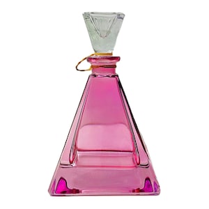 May include: A pink glass perfume bottle with a clear glass stopper. The bottle has a triangular shape with a flat base and a narrow neck. A gold string is tied around the neck. The stopper is a clear, inverted pyramid shape.