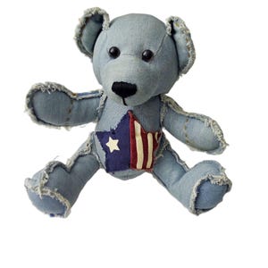 May include: A denim teddy bear with black button eyes and a black stitched nose. The bear has a star-shaped patch on its chest with a white star and red and white stripes. The edges of the denim are frayed.