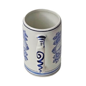 May include: A cylindrical ceramic beer stein featuring a white base with blue hand-painted floral patterns and landscape scenes. The handle is decorated with blue swirl motifs, and the rim and base are accented with thin blue horizontal lines.