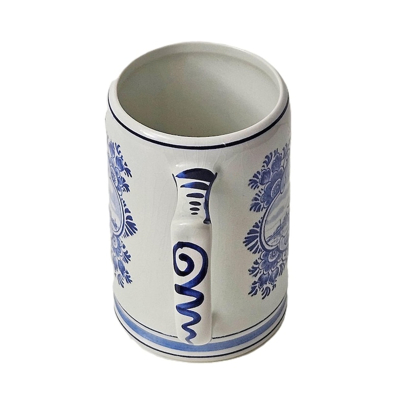 May include: A cylindrical ceramic beer stein featuring a white base with blue hand-painted floral patterns and landscape scenes. The handle is decorated with blue swirl motifs, and the rim and base are accented with thin blue horizontal lines.