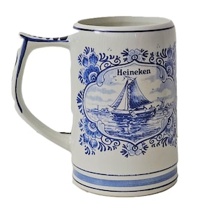 May include: A white ceramic beer mug featuring a blue decorative floral border and a central illustration of a sailboat on water. The word Heineken is printed in black above the sailboat. The handle and base are accented with blue stripes.