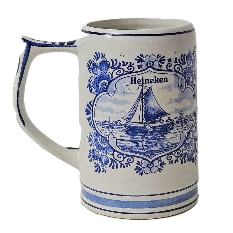 May include: A white ceramic beer mug featuring a blue decorative floral border and a central illustration of a sailboat on water. The word Heineken is printed in black above the sailboat. The handle and base are accented with blue stripes.