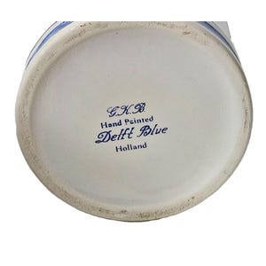 May include: The circular base of a ceramic object features blue text printed on a white background. The text reads G.K.B. Hand Painted Delft Blue Holland. A thin blue line circles the outer edge of the base, which has a slightly worn texture.