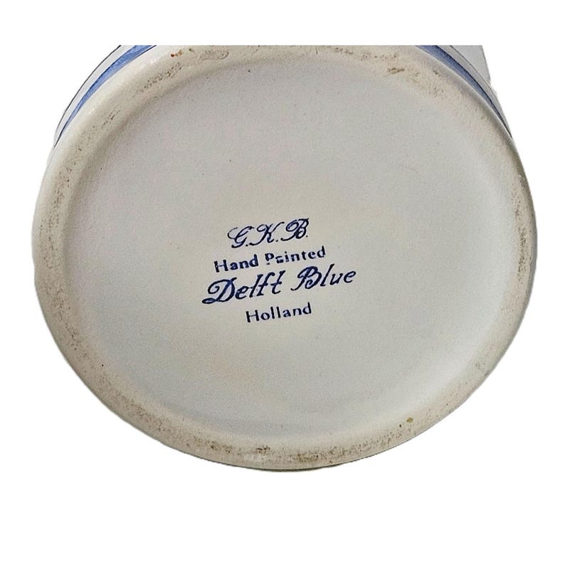 May include: The circular base of a ceramic object features blue text printed on a white background. The text reads G.K.B. Hand Painted Delft Blue Holland. A thin blue line circles the outer edge of the base, which has a slightly worn texture.