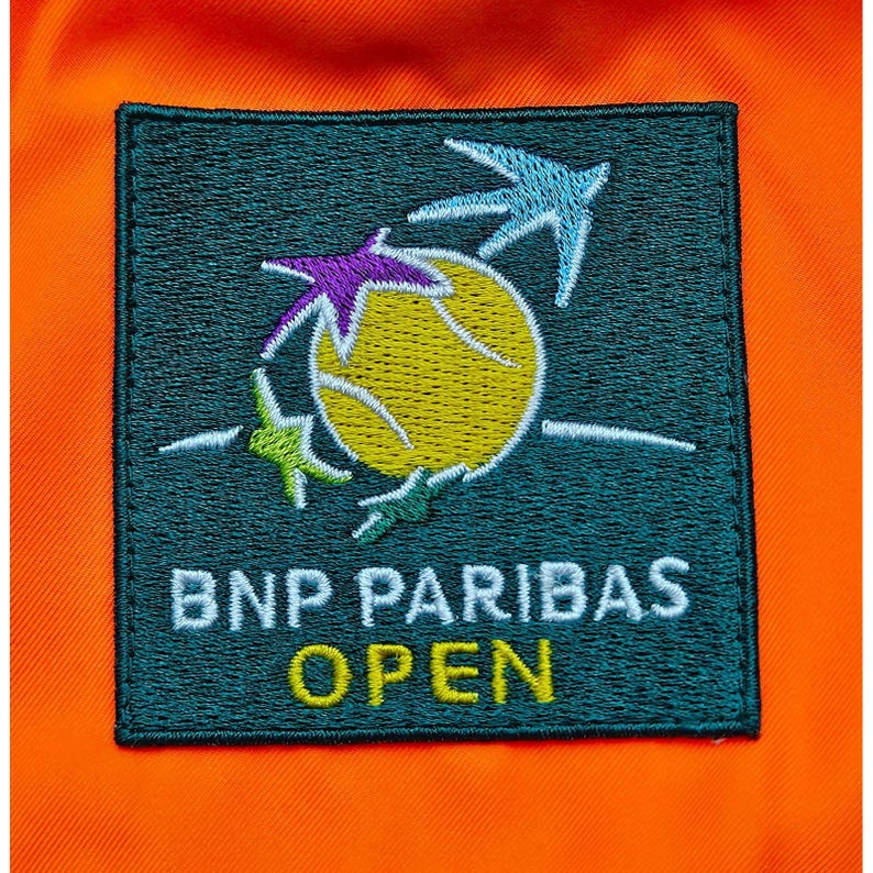 Peut inclure: &Eacute;cusson brod&eacute; avec le texte "BNP PARIBAS OPEN" en blanc et jaune. L'&eacute;cusson pr&eacute;sente un motif de balle de tennis jaune avec des accents en forme d'&eacute;toiles violettes, bleues et vertes. L'&eacute;cusson est sur un fond orange.